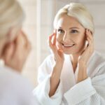 Smiling woman in a bathrobe applying skincare in front of a mirror