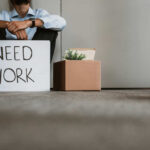 Person sitting on the ground holding a sign that says 'NEED WORK'