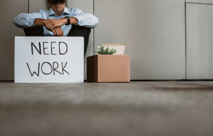 Person sitting on the ground holding a sign that says 'NEED WORK'