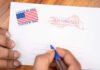A hand holding a pen writing on an official election mail envelope with a 'I Voted By Mail' sticker