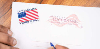 A hand holding a pen writing on an official election mail envelope with a 'I Voted By Mail' sticker