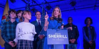 A woman in a blue dress speaks at a podium with supporters behind her