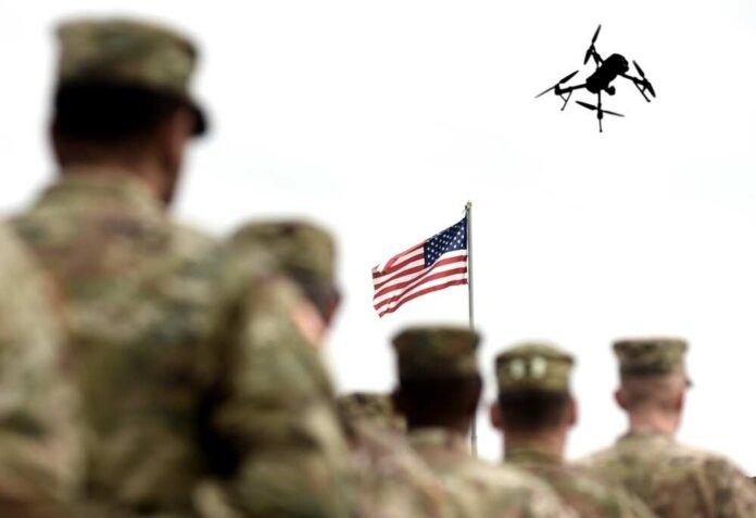 Military personnel standing in formation with a drone flying overhead and an American flag in the background
