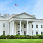 The White House with an American flag flying above, surrounded by greenery