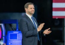 Man in a suit applauding at a political event with an American flag in the background