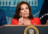 A woman in a red blazer speaking at a podium with the Presidential seal