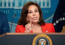 A woman in a red blazer speaking at a podium with the Presidential seal