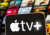 Close-up of a smartphone displaying the Apple TV+ logo with a blurred background of media thumbnails
