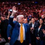 A man in a blue suit with a yellow tie waves to a cheering crowd