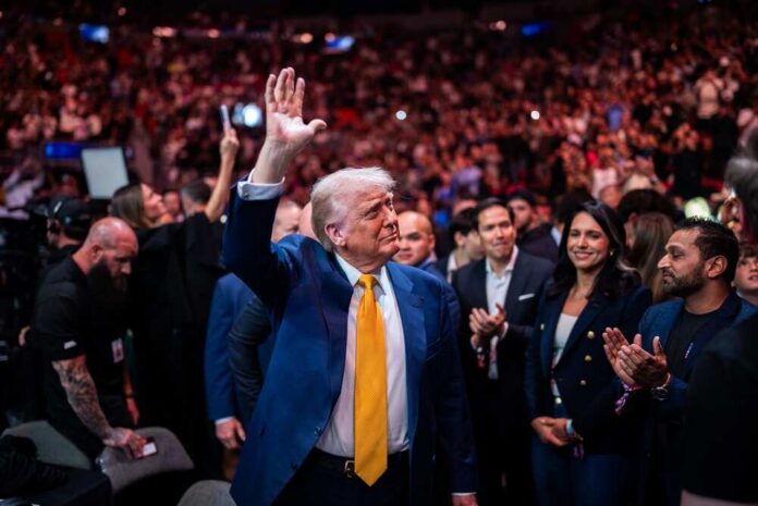 shutterstock_2747469381.jpg A man in a blue suit with a yellow tie waves to a cheering crowd