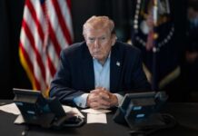 4% Drop: Trump Name Hurts Republicans? A serious-looking man seated at a table with phones and an American flag in the background