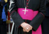 Chilling Threats Target Catholic School A bishop in traditional attire with a cross necklace and a pink sash, hands clasped in front