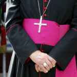 A bishop in traditional attire with a cross necklace and a pink sash, hands clasped in front