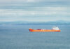 An orange cargo ship sailing in the ocean with a coastal landscape in the background