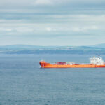 An orange cargo ship sailing in the ocean with a coastal landscape in the background