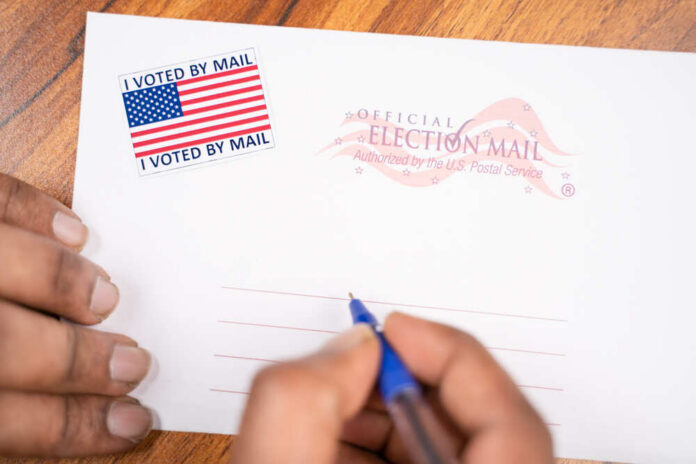 A hand holding a pen writing on an official election mail envelope with a 'I Voted By Mail' sticker