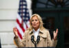 A woman speaking at a podium outdoors with an American flag in the background