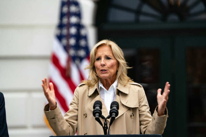 A woman speaking at a podium outdoors with an American flag in the background