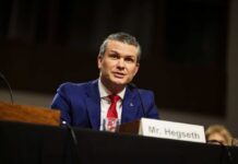 Hegseth Drops ‘Invasion’ Bomb at Normandy Man in a blue suit speaking at a government hearing