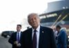 Trump’s Bold Iran Play Ignites G7 A political figure in a suit looking thoughtfully at an outdoor event with a ship in the background