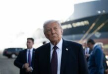 Trump’s Bold Iran Play Ignites G7 A political figure in a suit looking thoughtfully at an outdoor event with a ship in the background