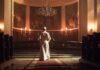 Schism Erupts: Bishops Banished Overnight A religious figure standing in a church with candlelight and historic artwork