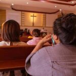 Church Sting Roils Talarico Bid people seated in church pews facing a cross