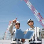 Two construction workers reviewing plans at a site with cranes