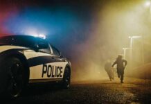 Police car with flashing lights at night as two people run in a foggy alley