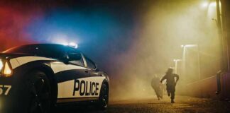 Police car with flashing lights at night as two people run in a foggy alley