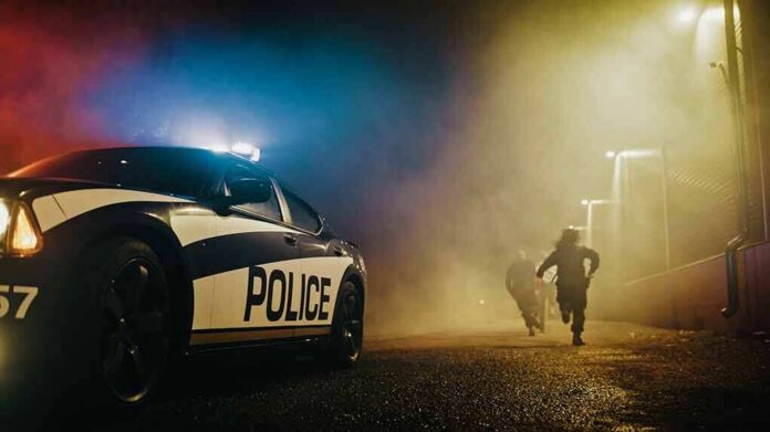 Police car with flashing lights at night as two people run in a foggy alley
