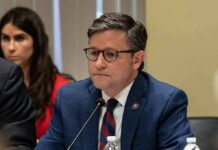 Impeach Fight Erupts – Speaker Pulls The Plug man in a blue suit speaking into a microphone at a meeting