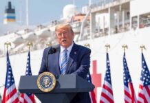 SHOCKING Memo Guts Endangered Protections man in suit speaking at a podium with flags and a ship behind
