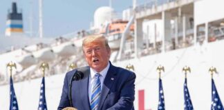 man in suit speaking at a podium with flags and a ship behind