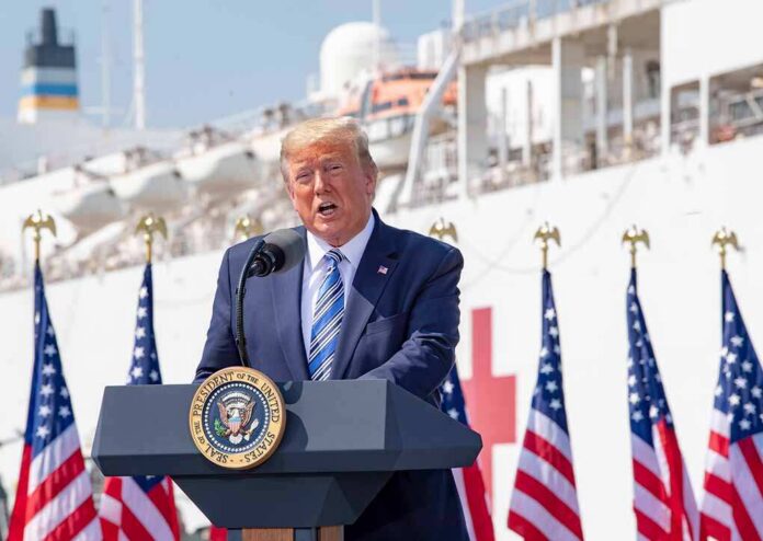 man in suit speaking at a podium with flags and a ship behind
